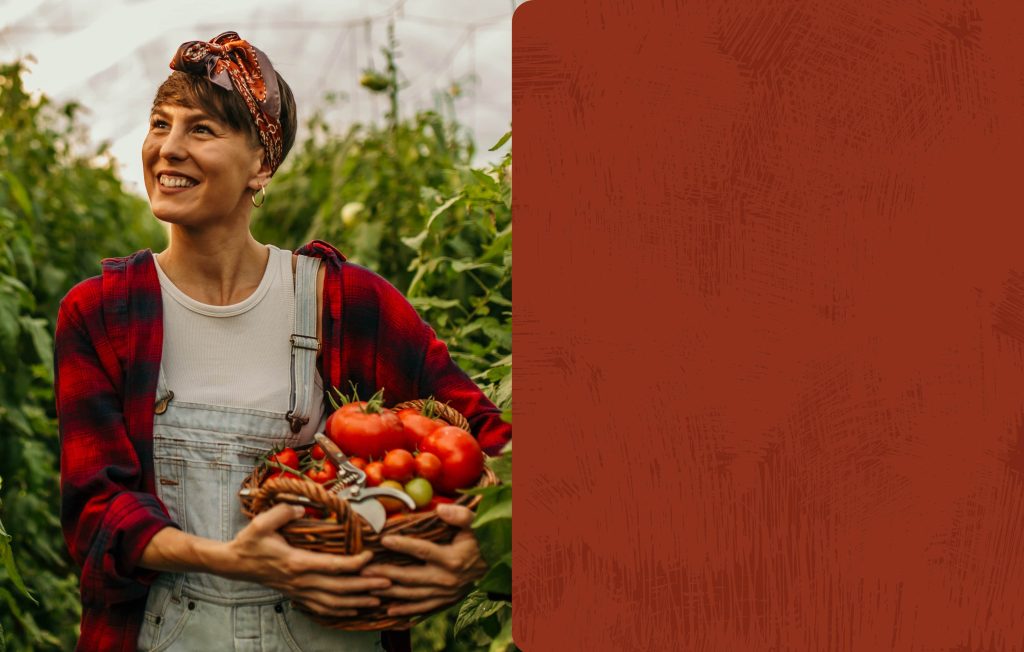 Smiling woman in denim overalls and a red plaid shirt holding a wicker basket full of freshly harvested tomatoes.